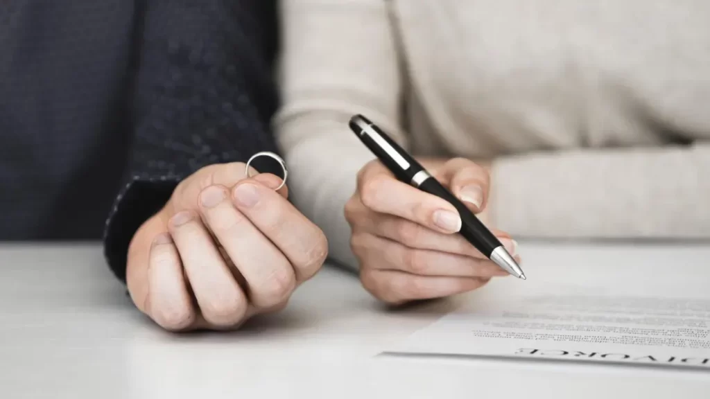 A husband sitting at a table writing a legal divorce notice for a NADRA certificate, while his wife stands nearby and hands her wedding ring back to him.